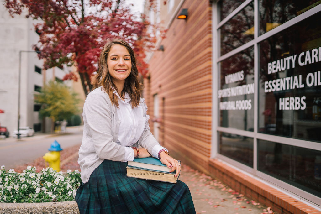 young woman smiling at camera while wearing plaid skirt and holding books after recording podcast episode about books your english teacher is obsessed with