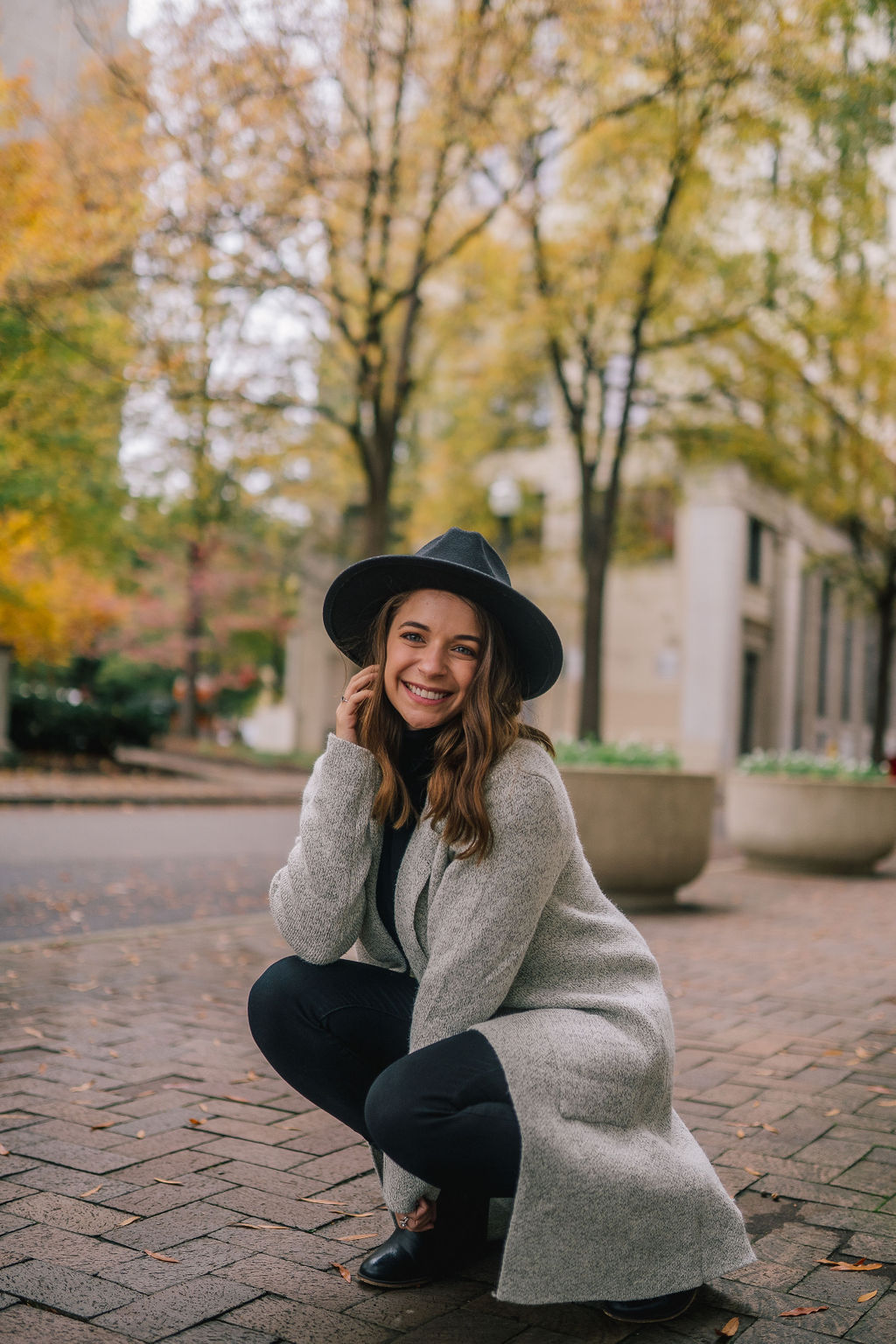 Anna kneeling and smiling wearing a long grey coat and black hat
