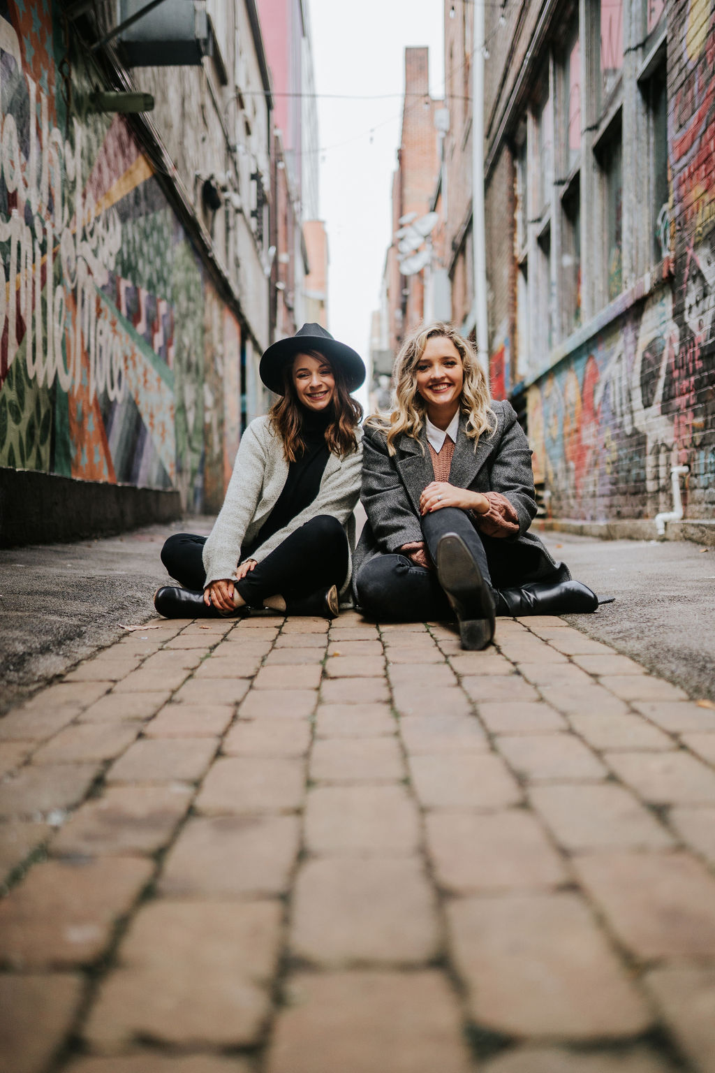 Celine and Anna from Novelbound a comedy book podcast sitting on cobblestone walkway in downtown knoxville laughing during novelbound podcast shoot